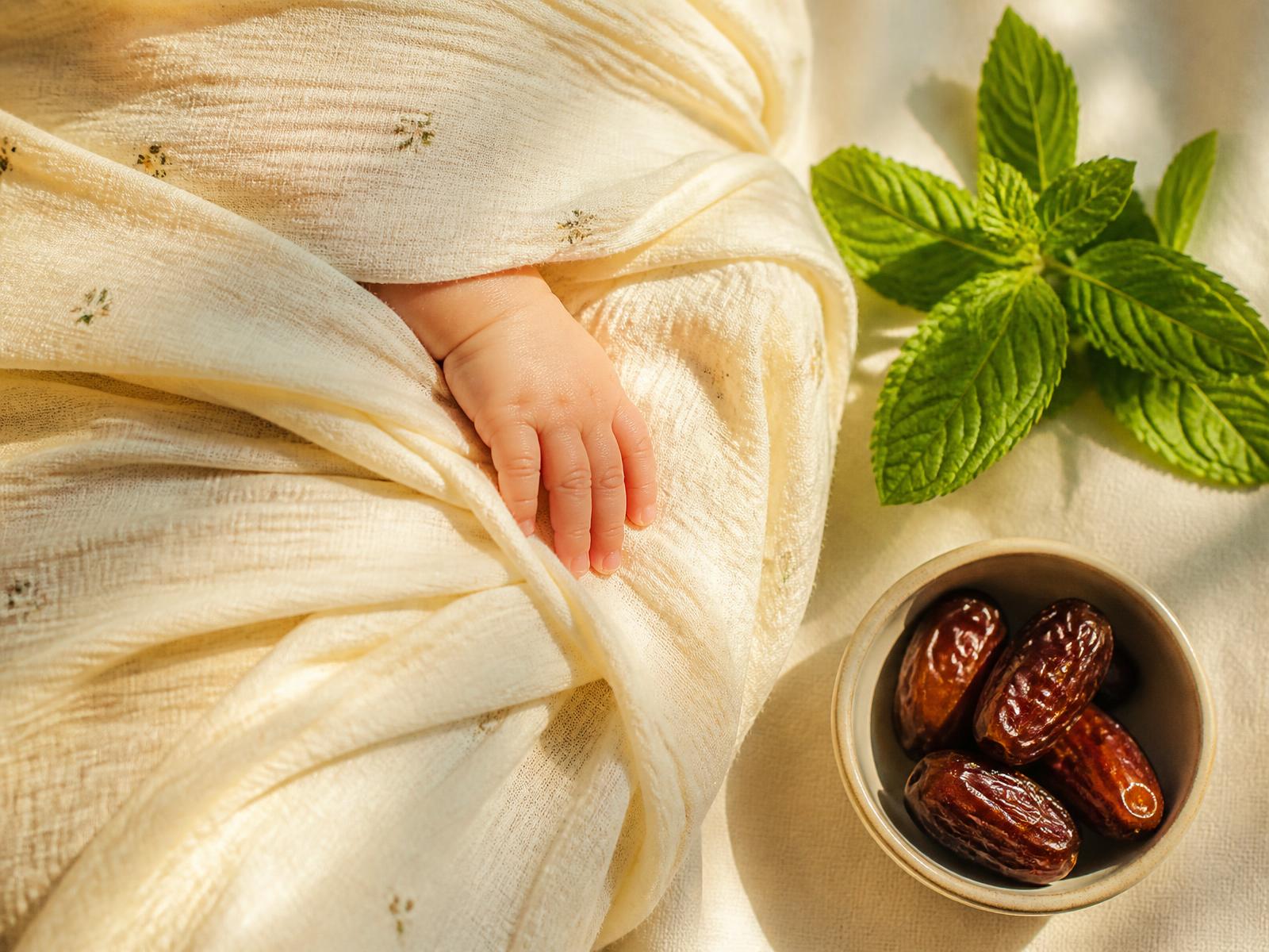 A sleeping newborn swaddled in cream muslin beside fresh mint and a bowl of dates